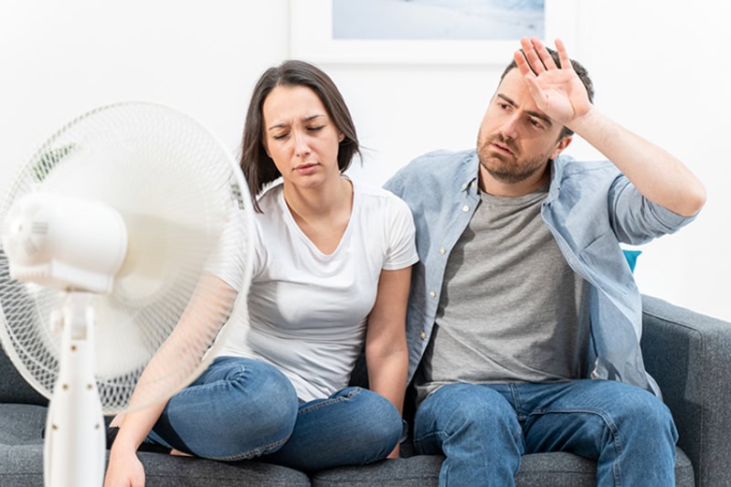 Why Is My AC Blowing Hot Air? An unhappy couple trying to cool off in their home in front of a fan.