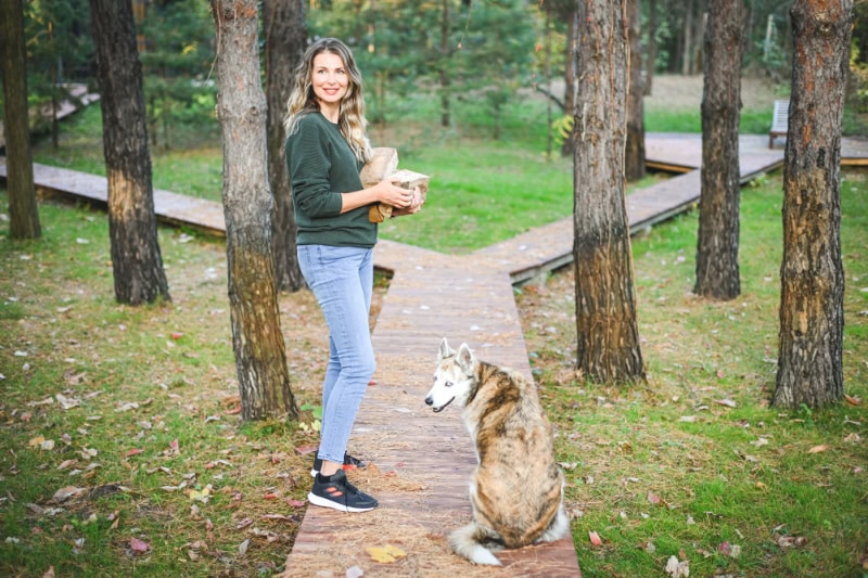 How Do I Know When It’s Time to Buy a New Furnace? Photo of a woman carrying firewood with her dog next to her on a wooden path into the woods.