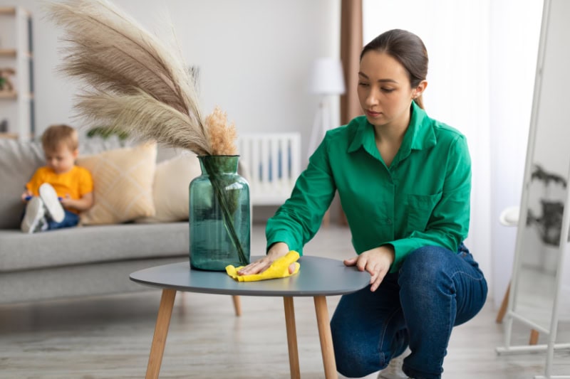Young mom dusting off coffee table and tidying up in living room while her toddler son watching cartoon on cellphone.